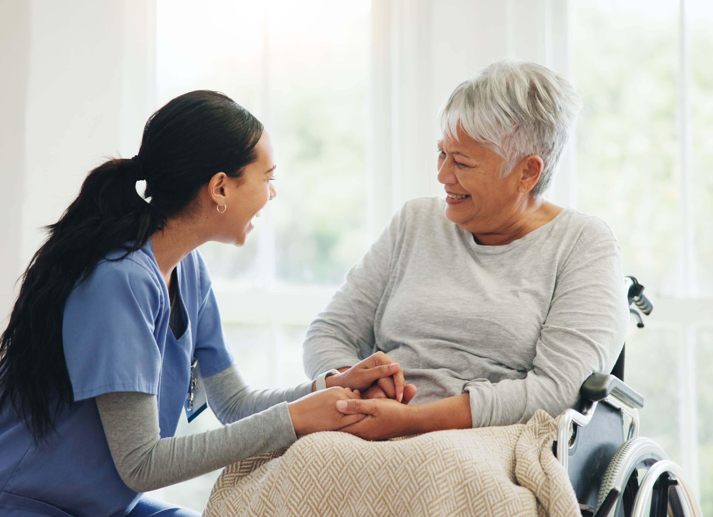 A nurse warmly holding hands with an elderly woman in a wheelchair.