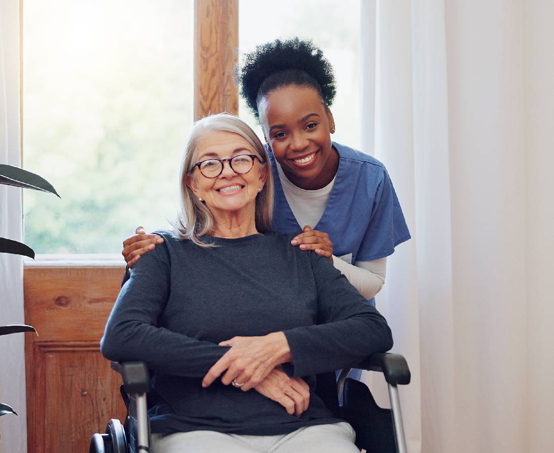 A caregiver embraces an elderly woman in a wheelchair, both smiling warmly.
