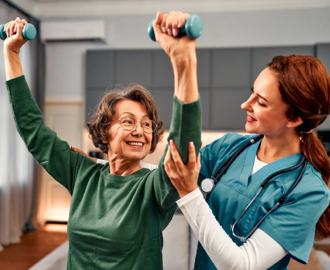 A nurse helps an elderly woman exercise with dumbbells, both smiling.