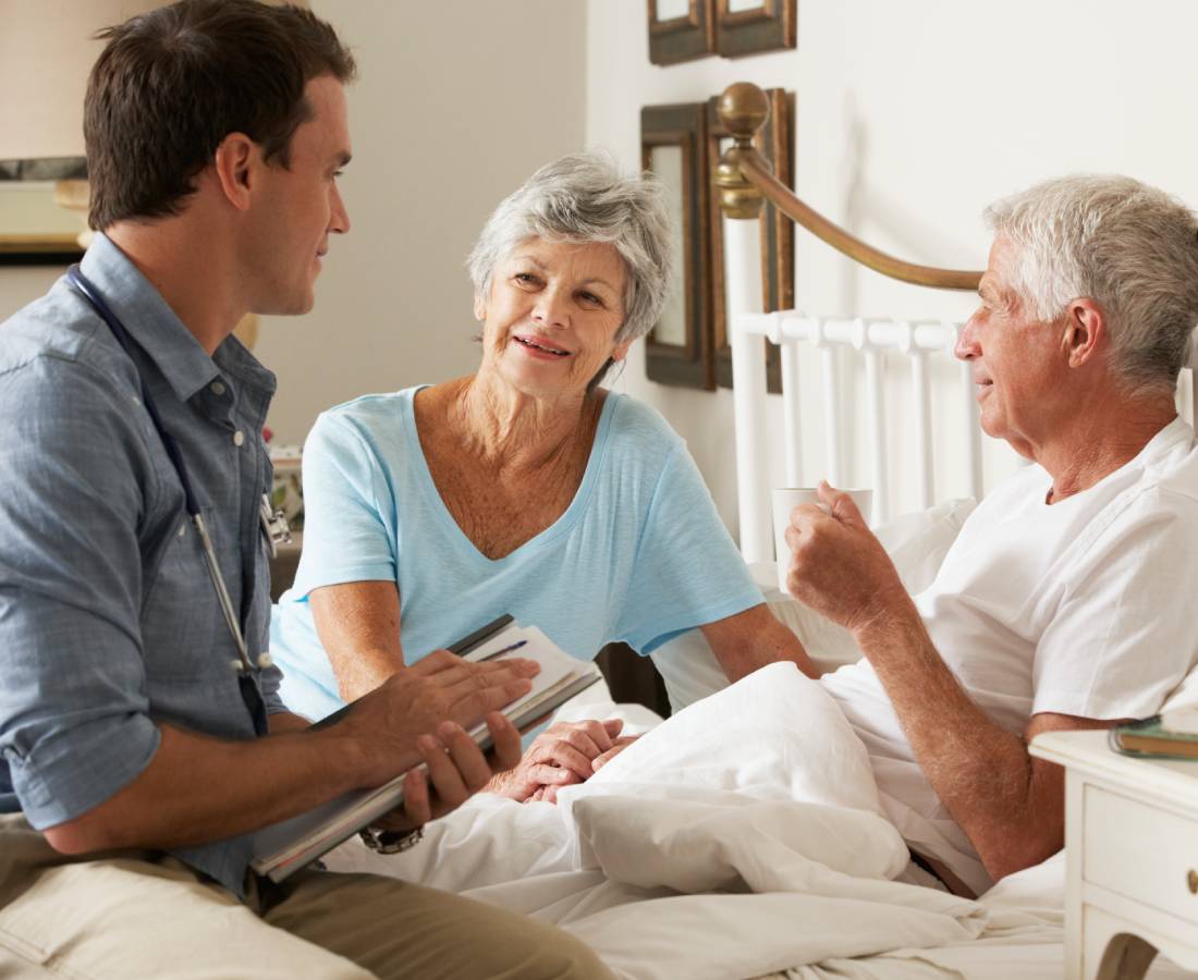 A young man talks with an elderly couple in bed, holding a tablet.