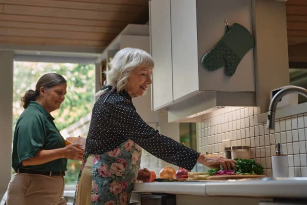 An elderly woman cooking in the kitchen with a younger person in the background.