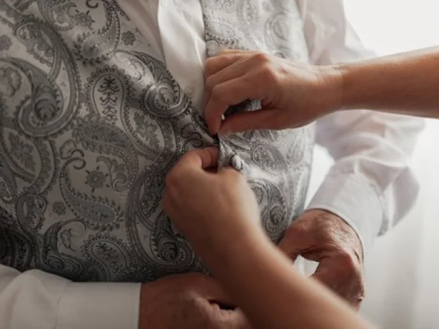 Hands buttoning a decorative vest on a person wearing a white shirt.