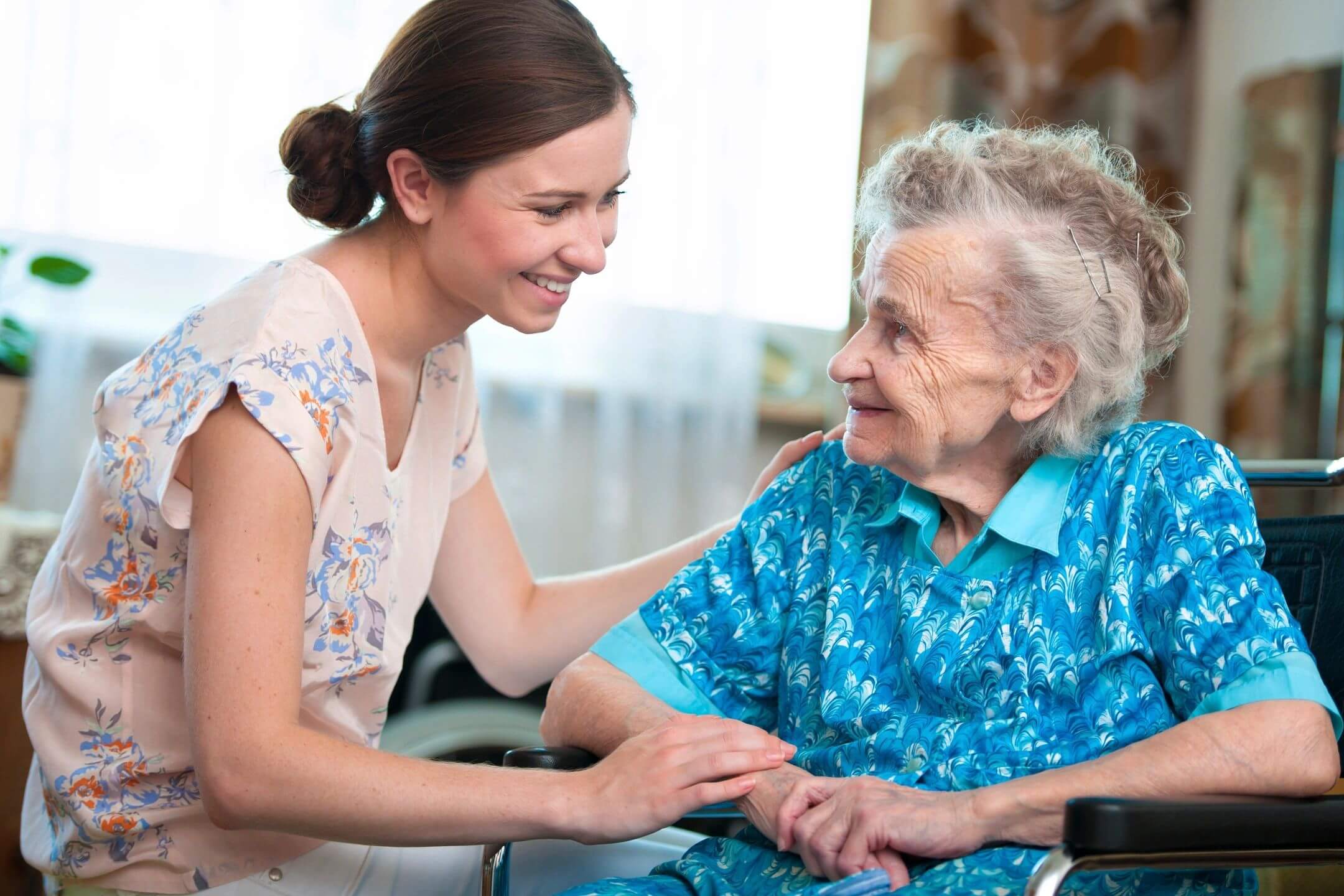 A young woman warmly holding hands with an elderly woman in a wheelchair.