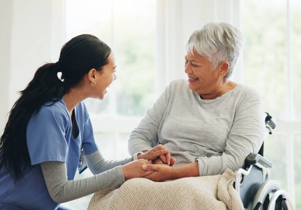 A nurse warmly holding hands with an elderly woman in a wheelchair.