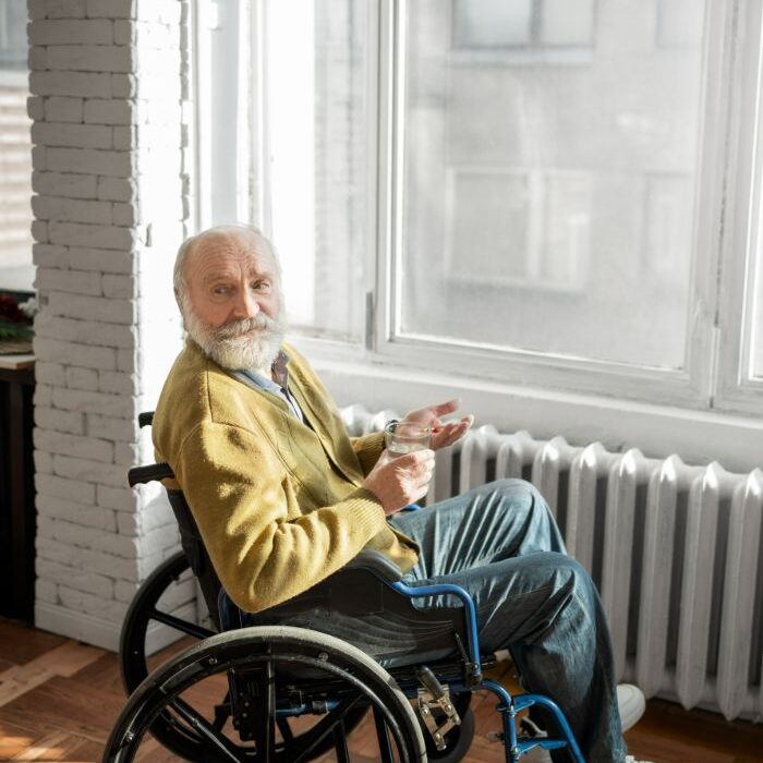 Elderly man in a wheelchair near a window holding a cup.